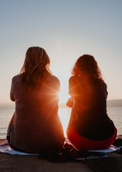 two women sitting on the beach watching the sunset and talking